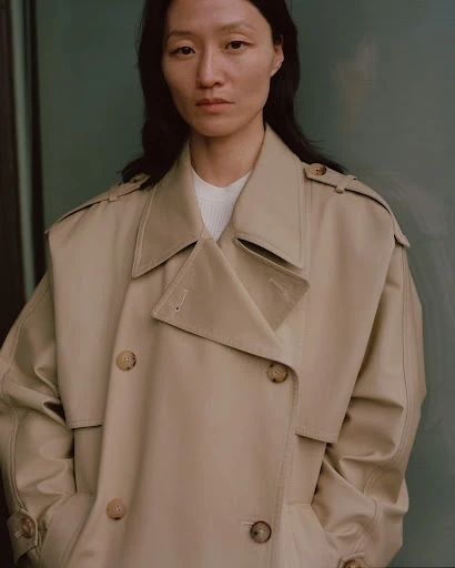 Portrait of a woman in a tan trench coat against a neutral green background, looking directly at the camera with a serene expression.