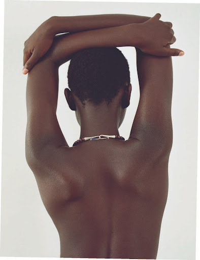 Back view of dark-skinned adult woman with arms crossed behind head and necklace, posing against plain light background.