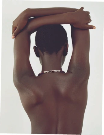 Back view of dark-skinned adult woman with arms crossed behind head and necklace, posing against plain light background.