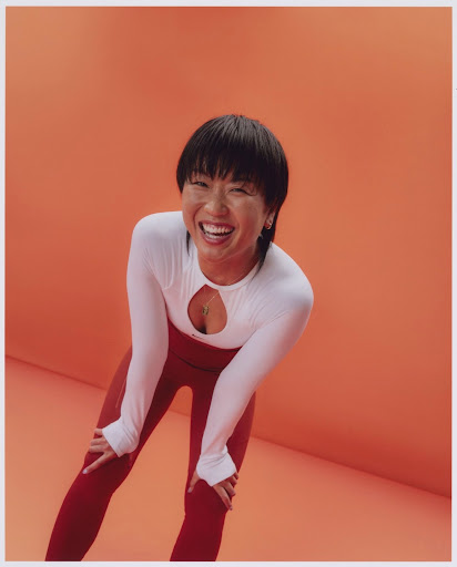 Smiling Asian woman in long-sleeved athletic top and leggings, leaning forward in an indoor fitness shot against an orange backdrop.