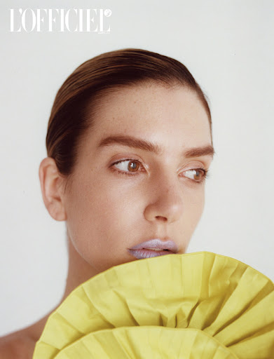 Close-up studio portrait of a woman with slicked-back hair and shimmery lilac lipstick, posing with a yellow ruffled garment.