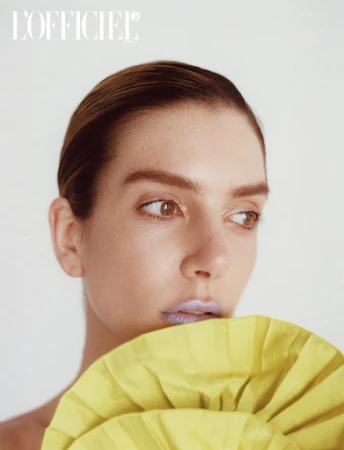 Close-up studio portrait of a woman with slicked-back hair and shimmery lilac lipstick, posing with a yellow ruffled garment.