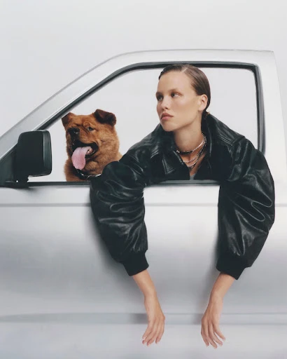 Adult woman in a black leather jacket with a Chow Chow dog in the silver truck leaning out of the window on a white background.
