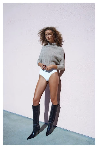 A woman poses against a pink wall, wearing a grey sweater, white bottoms, and black knee-high boots. Fashion editorial photography.