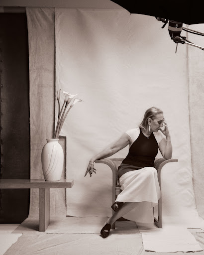 Elegant elderly woman in a black top and white skirt sits in a chair in a studio. Flowers and a softbox complete the portrait.