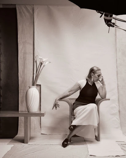 Elegant elderly woman in a black top and white skirt sits in a chair in a studio. Flowers and a softbox complete the portrait.