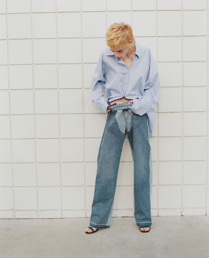 Woman in light blue button-down shirt and wide-leg denim pants poses against a textured white wall in a fashion shot.