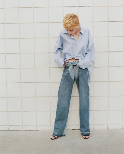 Woman in light blue button-down shirt and wide-leg denim pants poses against a textured white wall in a fashion shot.