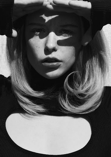 Black and white portrait of a young woman with hands raised, casting shadow on her face, wearing a black long-sleeved shirt and modern hairstyle.