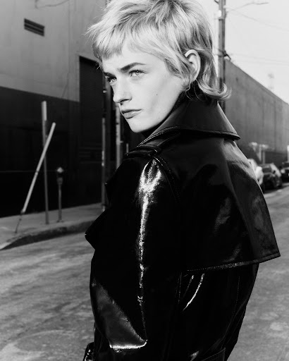 Monochrome portrait of a blonde adult female with short textured hair, wearing a shiny black coat and glancing to the left outdoors.