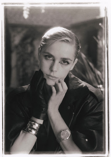 Monochrome portrait of a blonde adult woman wearing a leather jacket, metallic bracelets, and a watch, set against a blurred backdrop.