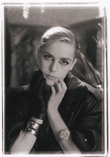 Monochrome portrait of a blonde adult woman wearing a leather jacket, metallic bracelets, and a watch, set against a blurred backdrop.