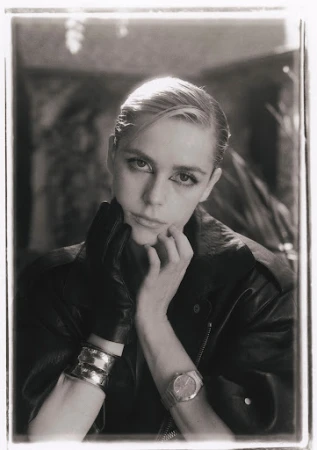 Monochrome portrait of a blonde adult woman wearing a leather jacket, metallic bracelets, and a watch, set against a blurred backdrop.