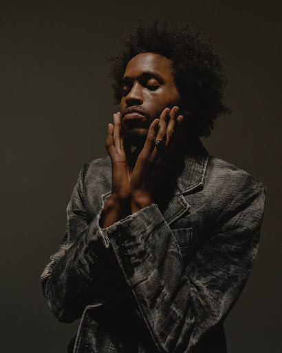 Portrait of a black man with an afro wearing a textured grey jacket with hands raised to his face.