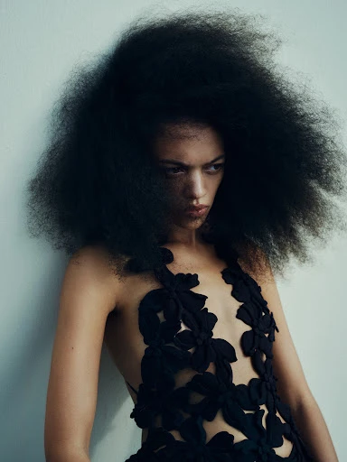 Adult woman with large Afro hairstyle wearing a black floral-patterned dress, posing in an indoor setting with soft, diffused lighting.