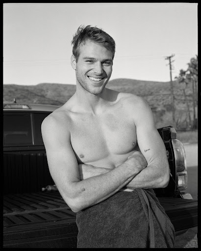 Black and white portrait of a smiling, shirtless man with arms crossed, wearing a towel, standing near a truck outdoors.