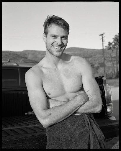 Black and white portrait of a smiling, shirtless man with arms crossed, wearing a towel, standing near a truck outdoors.