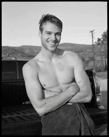 Black and white portrait of a smiling, shirtless man with arms crossed, wearing a towel, standing near a truck outdoors.