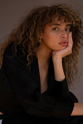 Portrait of a woman with curly brown hair resting her chin on her hand, wearing a black top against a solid neutral backdrop.