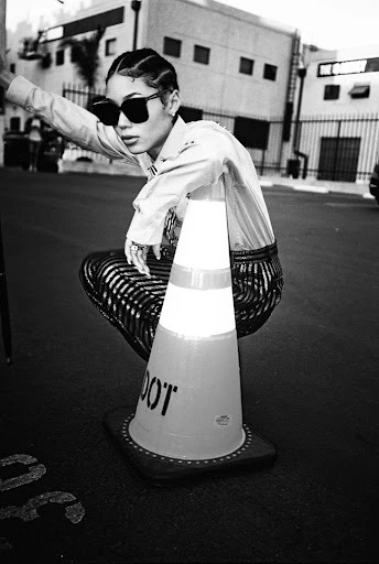 Black and white shot of a female fashion model posing next to a traffic cone on a city street with buildings in the background.