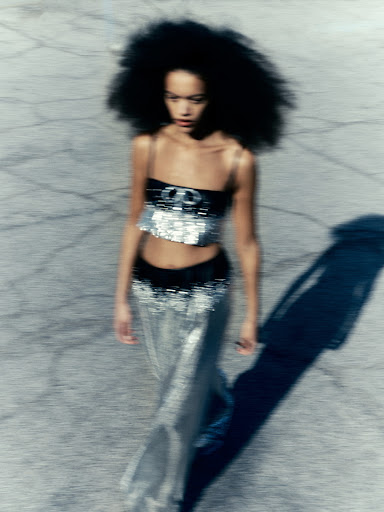 Fashion shot: a Black female model with a large afro poses outdoors in a silver sequin top and long skirt on cracked asphalt.
