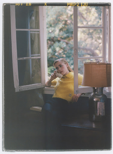 Woman in yellow shirt sits by window, talking on vintage telephone in a room with a table lamp and view of green outdoor trees.