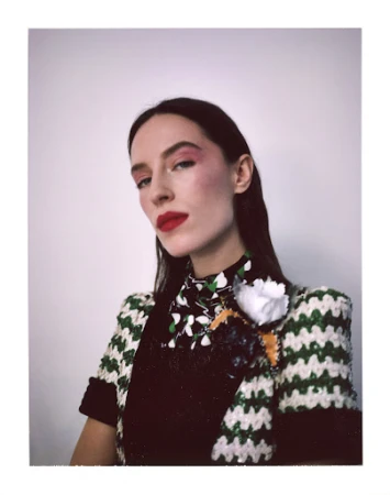Portrait of a woman wearing a green and white knitted top with a floral print collar and a white flower brooch against a white background.