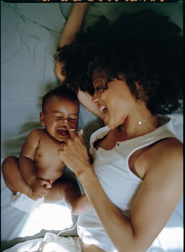 Mother and baby lying on a blue sheet, interacting with each other. The mother is smiling at the infant who is lying on his back.
