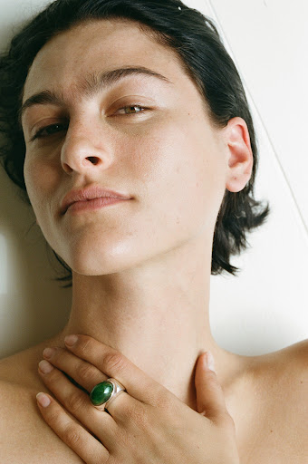Portrait of a young adult woman with dark hair and green ring, fair skin, and light make-up, looking calmly into the camera in soft lighting.