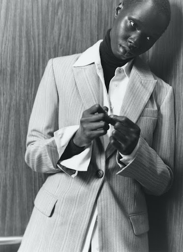 Black and white studio shot of a Black adult female wearing a suit, collared shirt, and turtleneck sweater, posing against a wood wall.