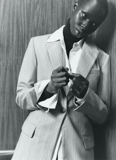 Black and white studio shot of a Black adult female wearing a suit, collared shirt, and turtleneck sweater, posing against a wood wall.