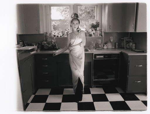 Monochrome photo of a woman in a dress standing in a kitchen with checkerboard floor, cabinets, and a window.