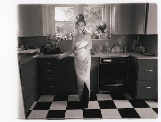 Monochrome photo of a woman in a dress standing in a kitchen with checkerboard floor, cabinets, and a window.