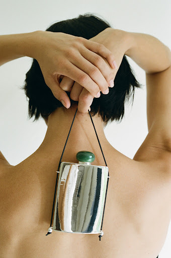 A woman is shown from the back, holding the straps of a flask with a green stopper. She has dark hair and is in front of a white background.