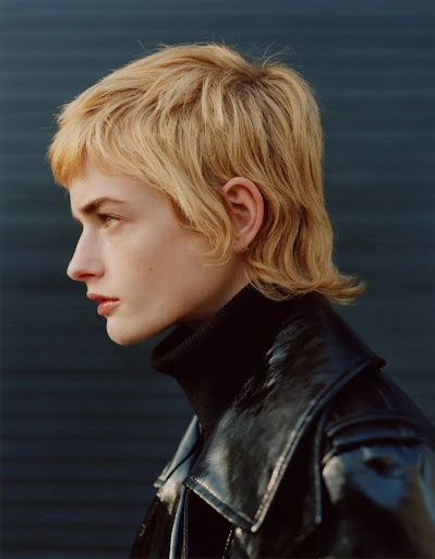 Profile of a blonde woman with a shag haircut wearing a black turtleneck and shiny jacket against a dark background in a studio shot.