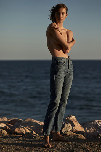 Topless woman wearing blue jeans standing on a rocky shore by the ocean at sunset with hair styled by Kelly Peach