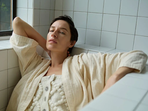 Woman with short dark hair in a cream textured cardigan leaning against white tiled walls