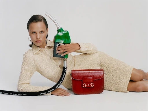 Adult female model in a beige textured dress lays next to a red purse and a green gas pump nozzle, all against a white backdrop.