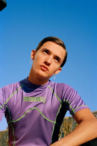 Young adult poses in a lavender athletic shirt against a clear blue sky, accessorized with hoop earrings. Outdoors, eye-level shot.