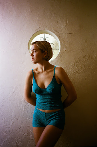 A woman in teal lingerie stands against a textured wall with a round window, bathed in soft, diffused light, creating a serene indoor portrait.