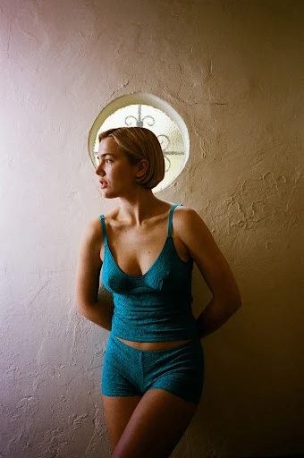 A woman in teal lingerie stands against a textured wall with a round window, bathed in soft, diffused light, creating a serene indoor portrait.