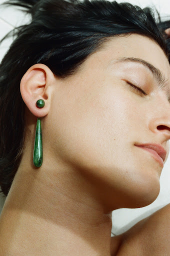 Close-up of a young adult woman with dark hair wearing green teardrop earrings against a plain light background with her eyes closed.