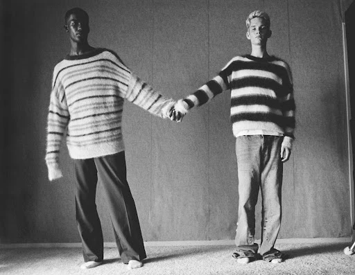 Two fashion models pose holding hands indoors wearing striped knit sweaters and slacks in a black and white editorial fashion shot.
