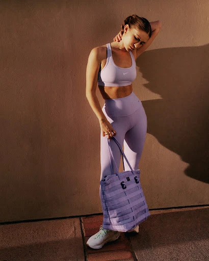 An adult woman stands outdoors in athletic wear with a purple tote bag. She is posing next to a textured wall with natural light.