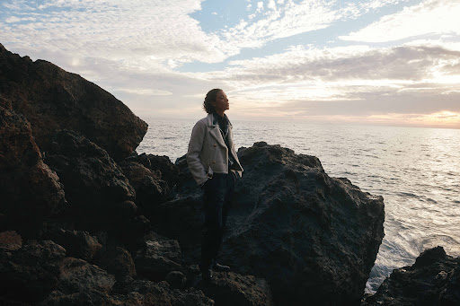 A person standing on dark coastal rocks by the ocean at sunset, wearing a light-colored jacket and dark trousers.