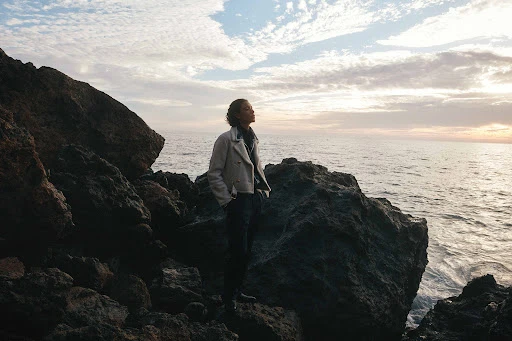 A person standing on dark coastal rocks by the ocean at sunset, wearing a light-colored jacket and dark trousers.