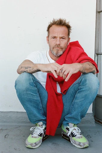Adult male with blonde hair sits in a crouched position wearing jeans, sneakers, and a white tee shirt. Red sweater draped over shoulders.