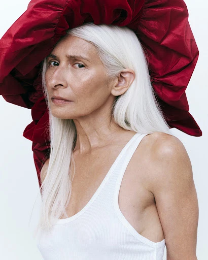 Close-up portrait of an older woman with long gray hair wearing a white tank top and a large, ruffled red hat against a white backdrop.