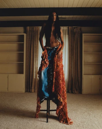 Model with long dark hair wrapped in a textured orange fringed blanket standing on a stool in a domestic interior.