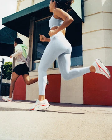 Woman in pale blue athletic wear running past a storefront with a man running in the background
